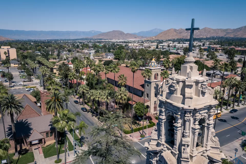 Aerial view of Riverside, California with ornate Catholic church tower in foreground and mountains in the distance.